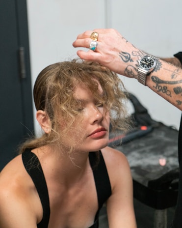 A woman with curly hair is receiving a hair touch-up from a person with tattooed arms and rings. The setting appears to be indoors, possibly a salon or dressing room, with a gray countertop visible in the background.
