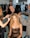 A group of stylists is working on preparing a model for a fashion event. The model is seated, wearing a black lace dress, while three stylists attend to her hair using tools such as a curling iron. The workspace is bustling with activity in the background, with racks of clothes and people moving around.