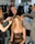 A group of stylists is working on preparing a model for a fashion event. The model is seated, wearing a black lace dress, while three stylists attend to her hair using tools such as a curling iron. The workspace is bustling with activity in the background, with racks of clothes and people moving around.