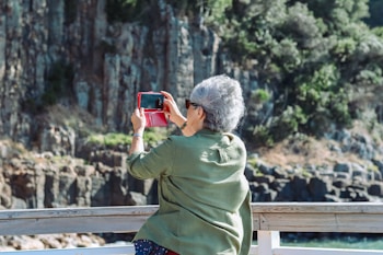 An older adult with short gray hair is standing on a wooden deck, holding a red smartphone case, taking a picture of a scenic outdoor view with rocky cliffs and greenery in the background. The person is wearing sunglasses and a green shirt.