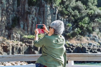 An older adult with short gray hair is standing on a wooden deck, holding a red smartphone case, taking a picture of a scenic outdoor view with rocky cliffs and greenery in the background. The person is wearing sunglasses and a green shirt.