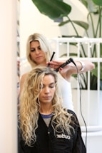 A hairstylist is curling the hair of a seated woman with a curling iron. The woman has long, wavy blond hair and is wearing a black salon cape. There is a plant in the background and a white railing behind the hairstylist.