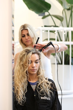 A hairstylist is curling the hair of a seated woman with a curling iron. The woman has long, wavy blond hair and is wearing a black salon cape. There is a plant in the background and a white railing behind the hairstylist.