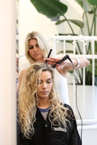 A hairstylist is curling the hair of a seated woman with a curling iron. The woman has long, wavy blond hair and is wearing a black salon cape. There is a plant in the background and a white railing behind the hairstylist.