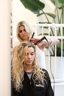 A hairstylist is curling the hair of a seated woman with a curling iron. The woman has long, wavy blond hair and is wearing a black salon cape. There is a plant in the background and a white railing behind the hairstylist.
