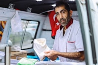A man stands inside a vehicle that is being used as a mobile food station. He is wearing a white uniform with red accents and holding a paper bag. The interior has a Turkish flag displayed on the wall.