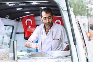 A man in a white shirt with red accents is serving food from a vehicle. Two Turkish flags are displayed in the background. The interior of the vehicle appears to be set up for food service, including a large pot and a towel.