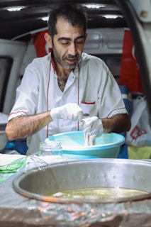 A man with short hair and a beard is preparing food in a mobile kitchen setup. He is wearing a white shirt with red accents and gloves while working with dough or a similar ingredient in a large blue bowl. In the foreground, there is a metal pan with hot oil, and various kitchen items scattered around.