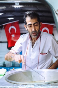 A man wearing a white shirt with red trim is preparing food, standing over a large metallic pot. Behind him, two Turkish flags are displayed, suggesting a cultural or national context. The setting appears to be a food stall or mobile kitchen.