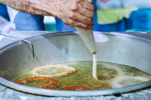 A hand pours batter into a large frying pan filled with hot oil, forming circular shapes as they fry. The scene captures the cooking process of a pastry or fritter, with one piece already golden brown while another one is being formed. The focus is on the circular formation and the hand holding a pastry bag, demonstrating a traditional cooking technique.