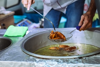 A person is using a metal strainer to lift pieces of food from a large pot filled with boiling oil. The food appears golden brown, suggesting it is being deep-fried. The surroundings include a counter covered with foil and a green cloth, and the person is wearing a casual shirt and jeans.