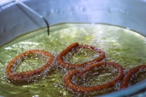 Golden brown rings of dough are frying in hot oil, with bubbles forming around them in the pan. The oil appears clear and the surface is reflective, highlighting the frying process.