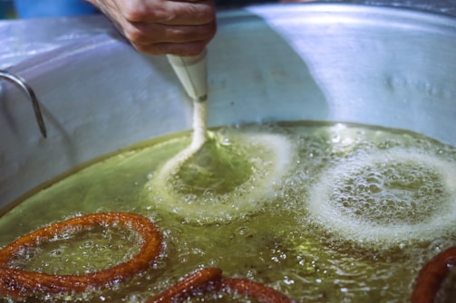 A person's hand is using a piping tool to create circular dough shapes in hot oil as they fry. The oil is bubbling around the dough, which is starting to turn golden brown.