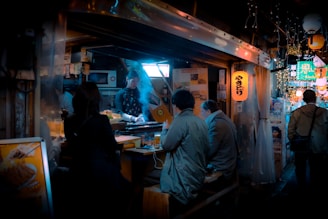 A small, cozy street food stall is bustling with activity. Several people are seated at the counter, while a chef is grilling yakitori skewers, with smoke rising in the air. The atmosphere is intimate and warm, with the soft glow of lanterns illuminating the space. Posters and various advertisements decorate the walls, contributing to the vibrant urban setting.