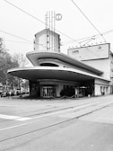 A modernist building with a distinctive, curved overhang and an Art Deco-inspired facade. The structure includes vertical signage with the words 'Garage Italia' and features a large front entrance. Surrounding the building are trees and a row of cars parked along the side. Tram lines and wires are visible overhead, indicating an urban setting.
