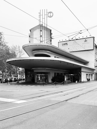 A modernist building with a distinctive, curved overhang and an Art Deco-inspired facade. The structure includes vertical signage with the words 'Garage Italia' and features a large front entrance. Surrounding the building are trees and a row of cars parked along the side. Tram lines and wires are visible overhead, indicating an urban setting.
