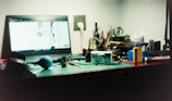 A cluttered desk with a laptop turned on, displaying a blurred, bright screen. Several items including a camera, a blue rubber bulb blower, film canisters, pens, and various small tools are scattered across the surface. The background features a wall outlet and partially visible writing instruments in a holder.