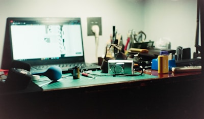 A cluttered desk with a laptop turned on, displaying a blurred, bright screen. Several items including a camera, a blue rubber bulb blower, film canisters, pens, and various small tools are scattered across the surface. The background features a wall outlet and partially visible writing instruments in a holder.