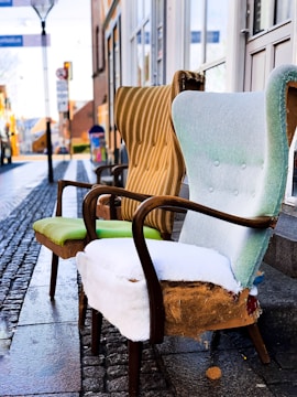 Two worn-out armchairs with elegant wooden frames are placed on a cobblestone street. The upholstery is old and torn, revealing padding beneath. Behind them, a blurred view of a street captures a few buildings and a lamppost, suggesting an outdoor urban setting.
