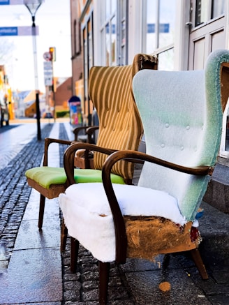 Two worn-out armchairs with elegant wooden frames are placed on a cobblestone street. The upholstery is old and torn, revealing padding beneath. Behind them, a blurred view of a street captures a few buildings and a lamppost, suggesting an outdoor urban setting.