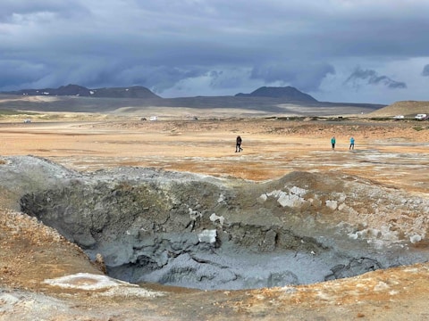 The image features a geothermal landscape with a large, cracked surface and a mud pool in the foreground. In the background, several people are walking across the barren terrain under a cloudy sky, with distant mountains and sparse vegetation.
