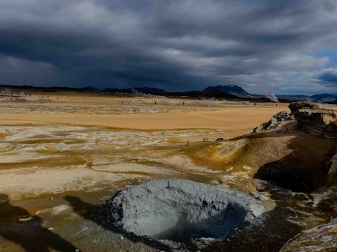 A vast geothermal landscape featuring a large, rocky geothermal vent in the foreground emitting steam. The ground is covered in various shades of yellow and white mineral deposits, while dark clouds loom overhead, casting dramatic shadows over the terrain. In the distance, small figures can be seen amidst more steaming vents, against a backdrop of distant mountains.
