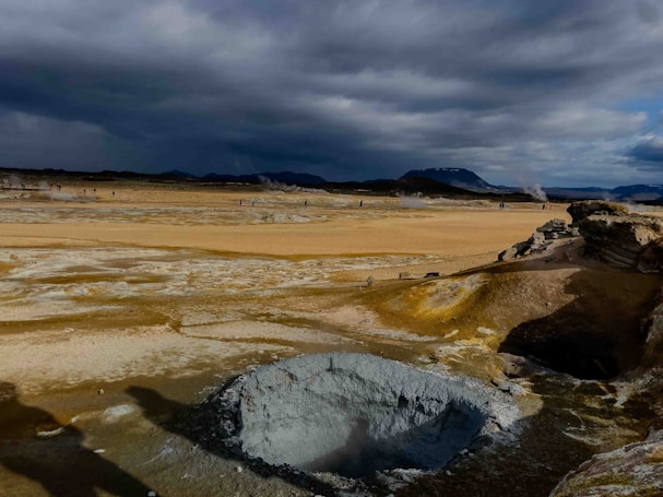 A vast geothermal landscape featuring a large, rocky geothermal vent in the foreground emitting steam. The ground is covered in various shades of yellow and white mineral deposits, while dark clouds loom overhead, casting dramatic shadows over the terrain. In the distance, small figures can be seen amidst more steaming vents, against a backdrop of distant mountains.