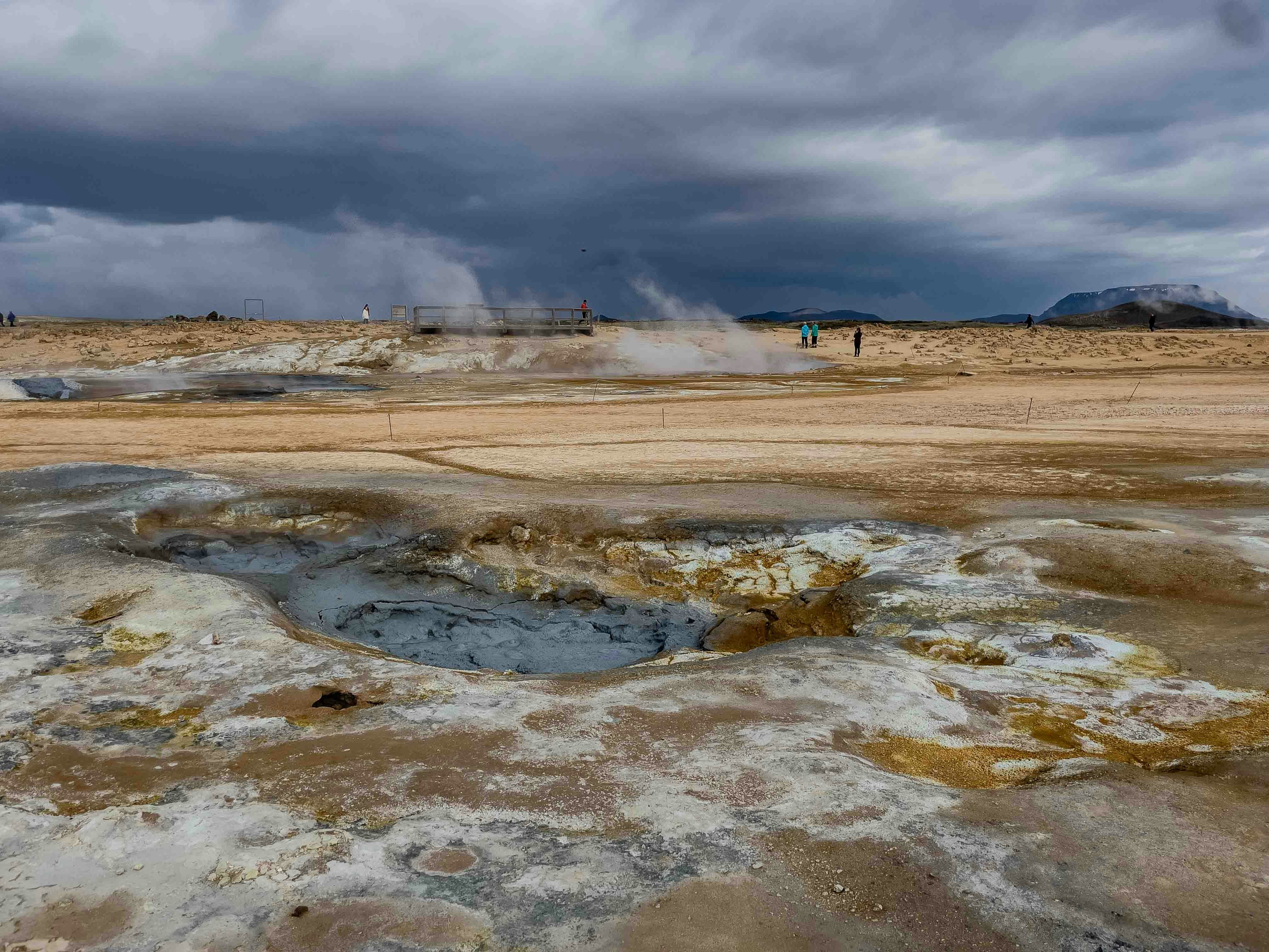 a group of people standing on top of a dry grass field, The image captures a group of tourists exploring a geothermal area with boiling mud pools and steam vents, set against a backdrop of a dramatic sky and distant mountains.