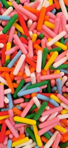 A vibrant assortment of colorful, cylindrical candy pieces scattered randomly. The candy pieces are predominantly pink, red, green, yellow, blue, and orange, creating a lively and playful visual effect.