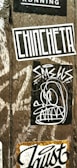 A wooden surface is covered with multiple street art stickers. One has the word 'CHINCHETA' in large, bold letters. Another features a cartoonish, abstract face with exaggerated eyes and teeth. Additional stickers include various text and graphic designs.