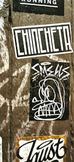 A wooden surface is covered with multiple street art stickers. One has the word 'CHINCHETA' in large, bold letters. Another features a cartoonish, abstract face with exaggerated eyes and teeth. Additional stickers include various text and graphic designs.