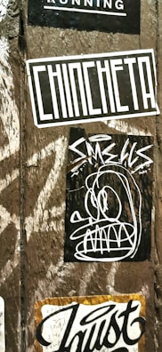 A wooden surface is covered with multiple street art stickers. One has the word 'CHINCHETA' in large, bold letters. Another features a cartoonish, abstract face with exaggerated eyes and teeth. Additional stickers include various text and graphic designs.