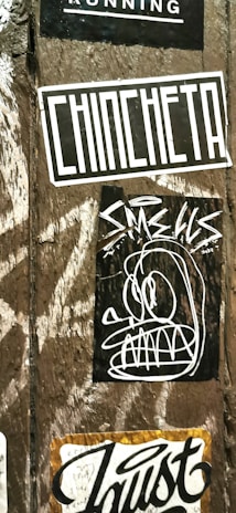 A wooden surface is covered with multiple street art stickers. One has the word 'CHINCHETA' in large, bold letters. Another features a cartoonish, abstract face with exaggerated eyes and teeth. Additional stickers include various text and graphic designs.