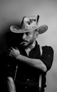 A person with a beard, wearing a wide-brimmed cowboy hat decorated with stitching and feathers. They have an earring and are dressed in a dark shirt with suspenders. The image is in black and white, creating a dramatic contrast.