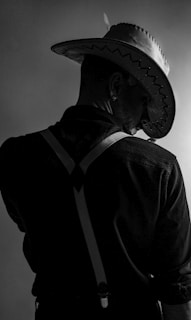 A person wearing a cowboy hat is seen from behind, with suspenders visible over a dark shirt. The lighting creates a dramatic shadow effect, emphasizing the texture of the hat and clothing.