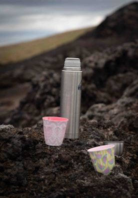 A metal thermos and three colorful cups are placed on a rocky surface in an outdoor setting. The background is blurred, showing muted landscapes and a cloudy sky. The cups feature vibrant patterns with pink and green colors.