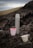 A metal thermos and three colorful cups are placed on a rocky surface in an outdoor setting. The background is blurred, showing muted landscapes and a cloudy sky. The cups feature vibrant patterns with pink and green colors.