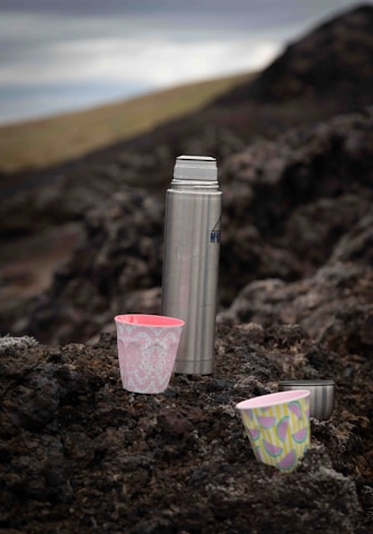 A metal thermos and three colorful cups are placed on a rocky surface in an outdoor setting. The background is blurred, showing muted landscapes and a cloudy sky. The cups feature vibrant patterns with pink and green colors.
