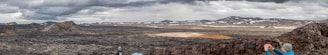 A vast, rugged landscape with rocky terrain and sparse patches of vegetation under an overcast sky. Mountains and hills are visible in the distance, with snow scattered across the elevated areas. In the foreground, a couple of people are taking photographs, capturing the dramatic scenery.
