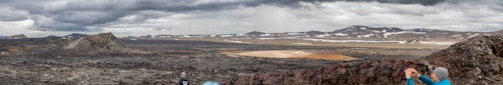 A vast, rugged landscape with rocky terrain and sparse patches of vegetation under an overcast sky. Mountains and hills are visible in the distance, with snow scattered across the elevated areas. In the foreground, a couple of people are taking photographs, capturing the dramatic scenery.