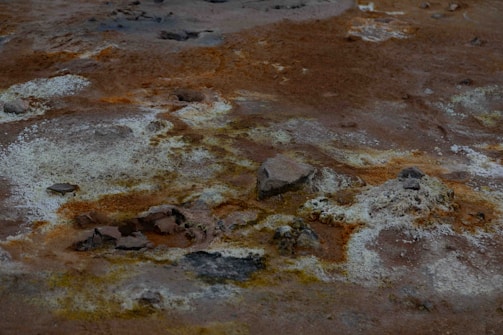 A barren landscape with a rugged surface, featuring patches of white, gray, and rusty orange hues. The ground is uneven, with rocky textures and small stones scattered across the area, indicating a dry, desolate environment.