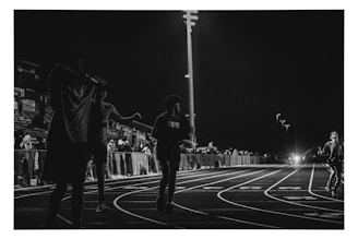 A black and white photograph captures a nighttime scene at a track and field event. Several figures are visible on the track, with one person running towards the camera. The background features a crowd of spectators sitting in the bleachers, illuminated by stadium lights.
