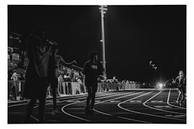 A black and white photograph captures a nighttime scene at a track and field event. Several figures are visible on the track, with one person running towards the camera. The background features a crowd of spectators sitting in the bleachers, illuminated by stadium lights.