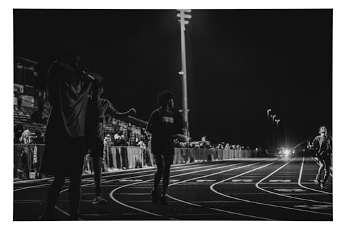 A black and white photograph captures a nighttime scene at a track and field event. Several figures are visible on the track, with one person running towards the camera. The background features a crowd of spectators sitting in the bleachers, illuminated by stadium lights.