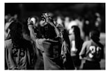 A black and white image featuring people wearing numbered sports jerseys. The focus is on individuals with numbers 28, 5, 11, and 39 visible on their jerseys. Some people have their hands raised and appear to be interacting in a communal or celebratory manner. The background is blurred, indicating others are present but out of focus.