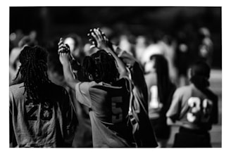 A black and white image featuring people wearing numbered sports jerseys. The focus is on individuals with numbers 28, 5, 11, and 39 visible on their jerseys. Some people have their hands raised and appear to be interacting in a communal or celebratory manner. The background is blurred, indicating others are present but out of focus.