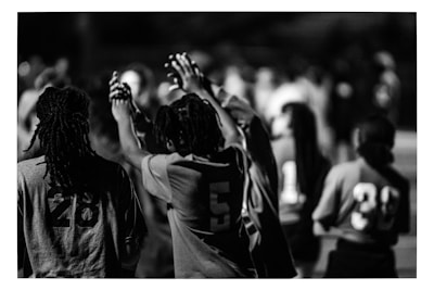 A black and white image featuring people wearing numbered sports jerseys. The focus is on individuals with numbers 28, 5, 11, and 39 visible on their jerseys. Some people have their hands raised and appear to be interacting in a communal or celebratory manner. The background is blurred, indicating others are present but out of focus.