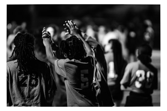 A black and white image featuring people wearing numbered sports jerseys. The focus is on individuals with numbers 28, 5, 11, and 39 visible on their jerseys. Some people have their hands raised and appear to be interacting in a communal or celebratory manner. The background is blurred, indicating others are present but out of focus.