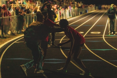 A nighttime scene at a track and field event with spectators in the stands. People are on the track, and one person appears to be assisting or supporting another. The environment is dark, with artificial lighting illuminating certain areas.