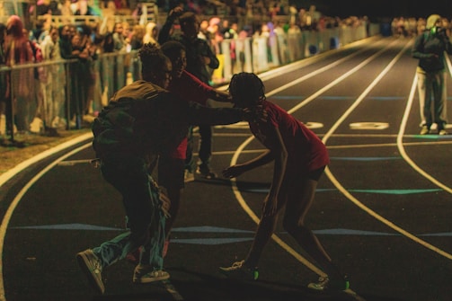 A nighttime scene at a track and field event with spectators in the stands. People are on the track, and one person appears to be assisting or supporting another. The environment is dark, with artificial lighting illuminating certain areas.